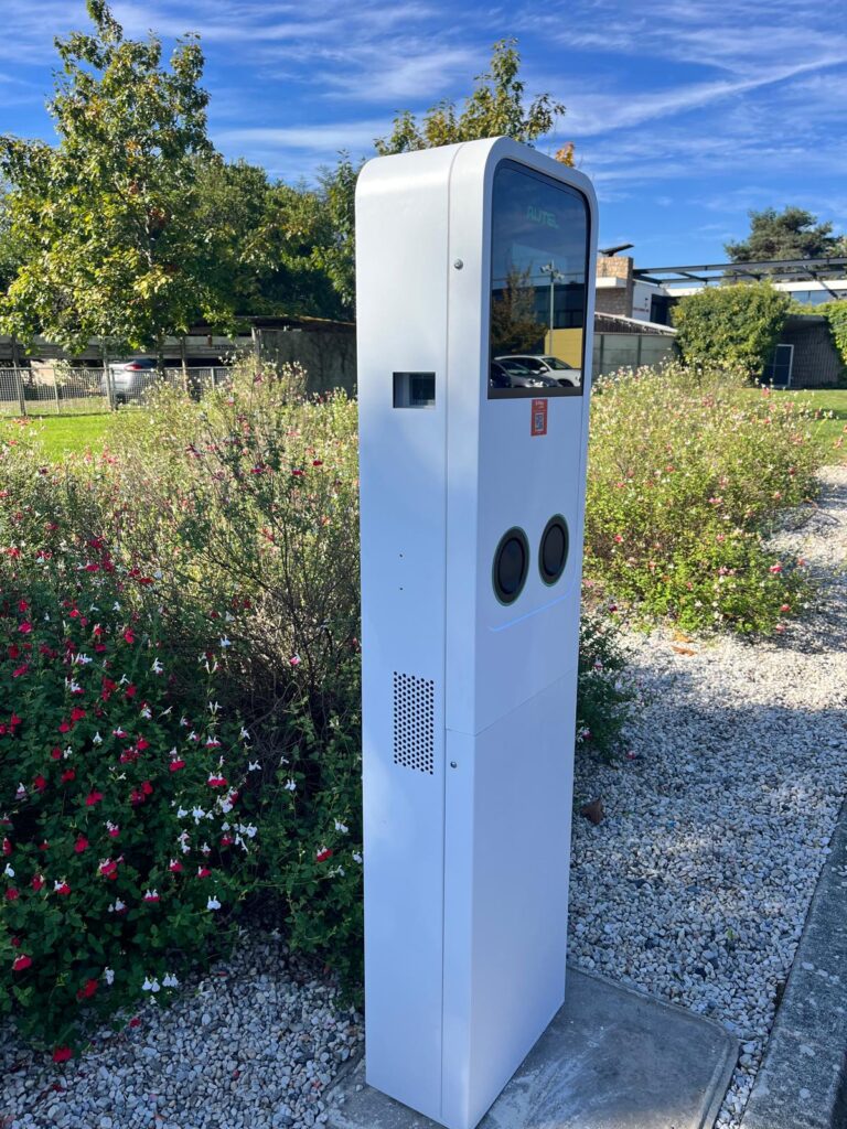 Borne de recharge électrique AUTEL blanche, moderne, entourée de fleurs rouges et blanches, avec un écran affichant 'AUTEL'.