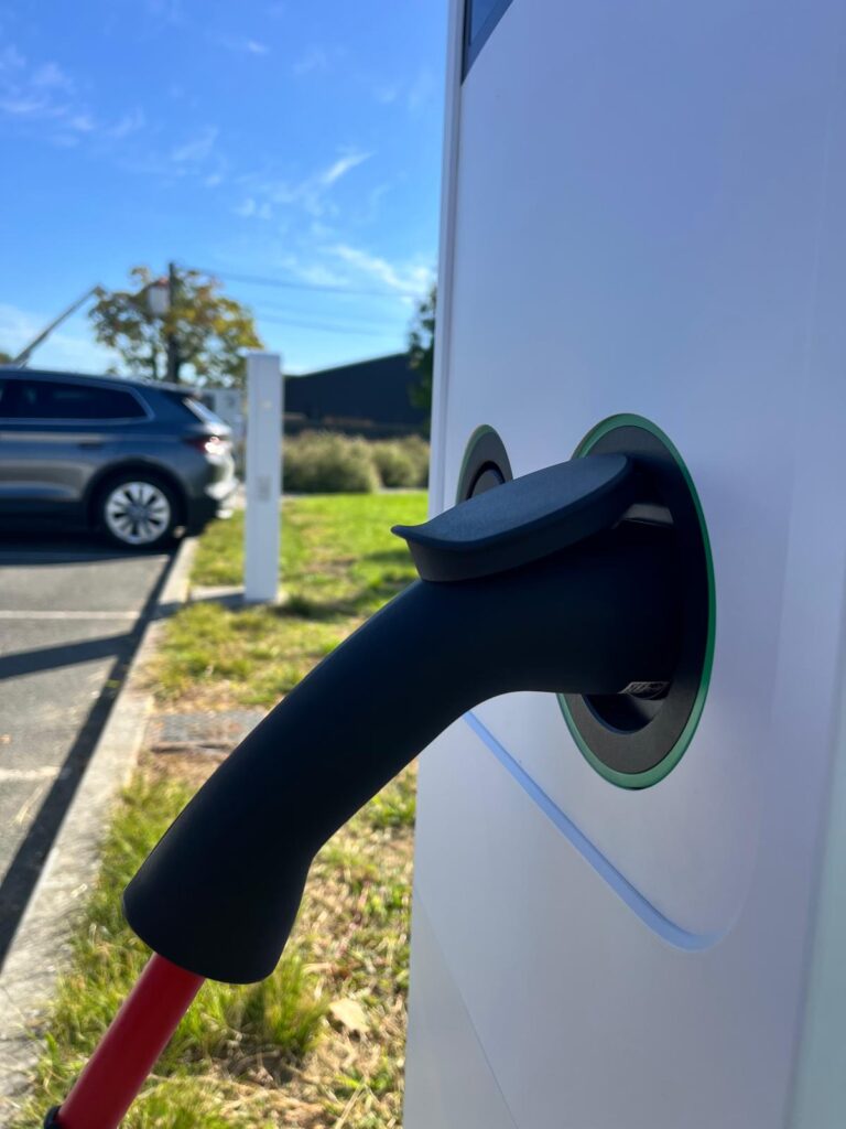 Gros plan sur une borne de recharge pour véhicule électrique avec le connecteur noir inséré, en extérieur, ciel bleu.